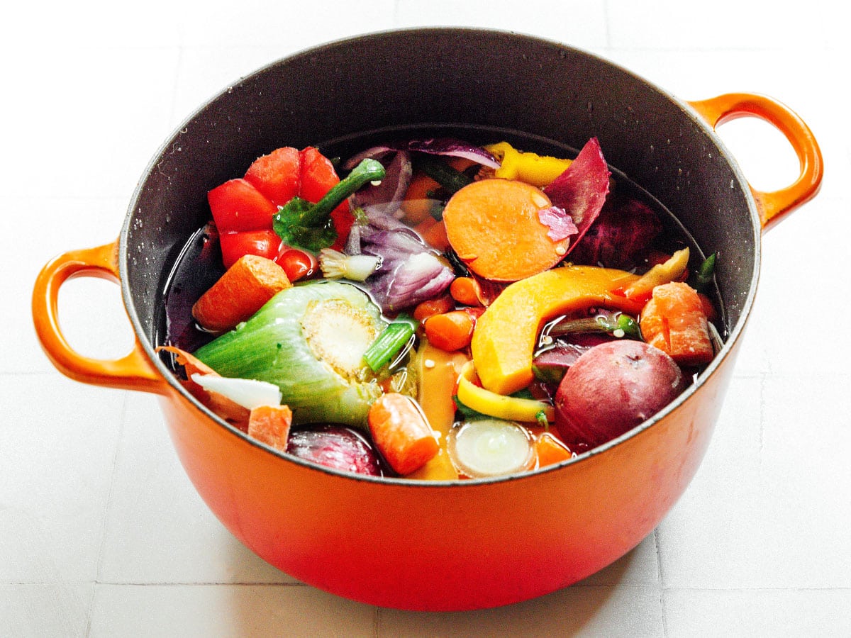Vegetable scraps including celery, bell pepper, butternut squash, potatoes, and carrots in a large orange pot with water. 