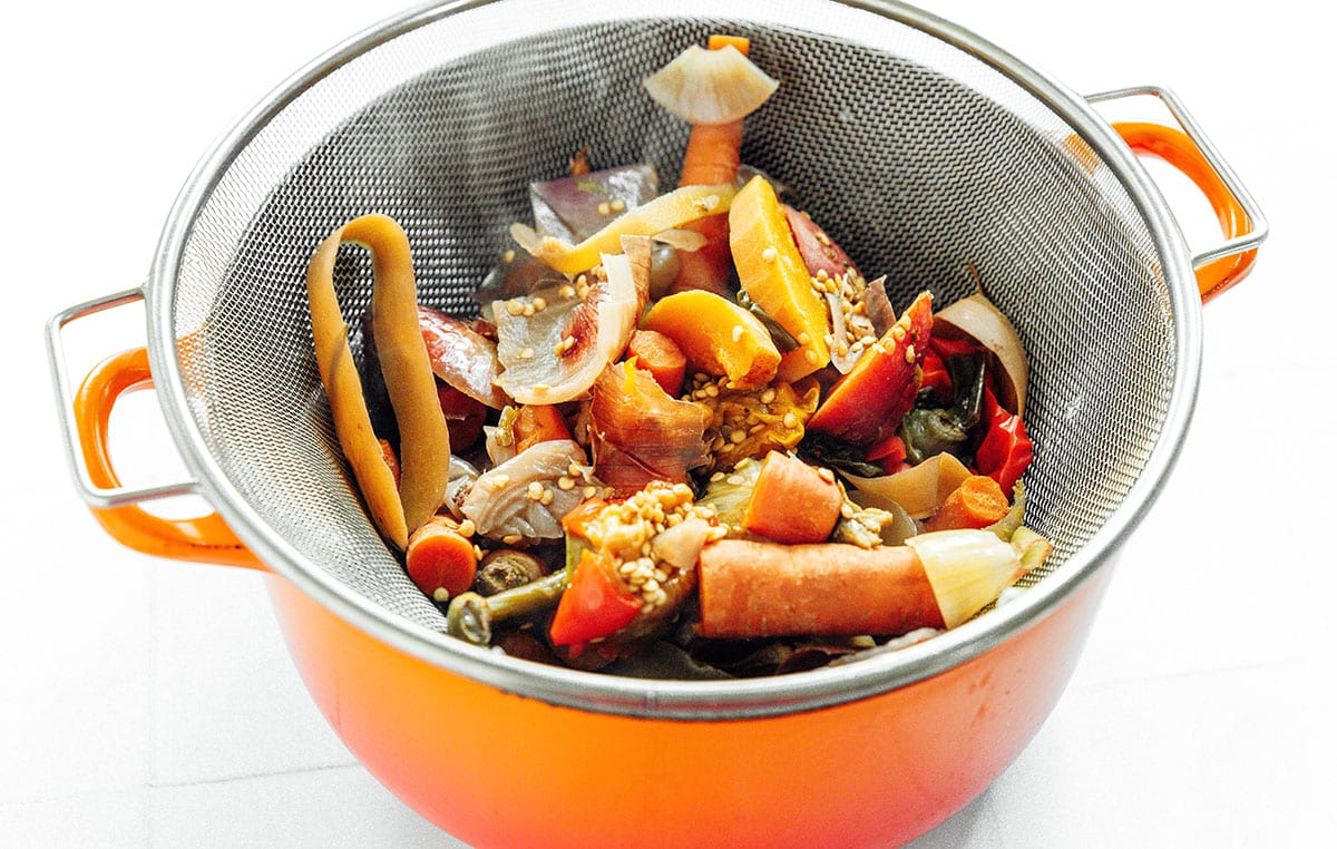 Cooked vegetable scraps in a large silver strainer, set on top of an orange pot. White background.