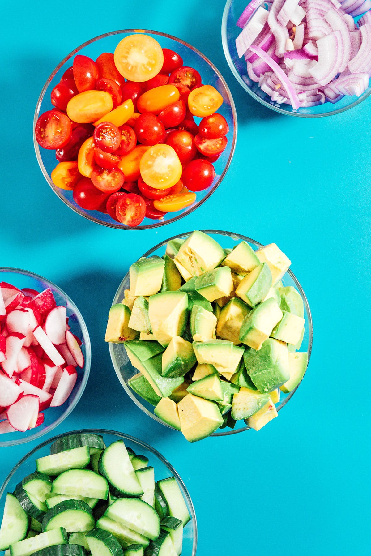 Separate bowls of bite-sized avocados, cherry tomatoes, and radishes sit on a blue background. The bowls are clear glass.