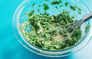 A glass bowl containing chopped herbs, minced garlic, and a liquid mixture for avocado salad with a metal whisk, set on a bright blue surface.