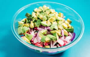 A glass bowl containing a vibrant avocado salad with diced avocado, sliced red onions, and fresh radishes on a bright blue surface.