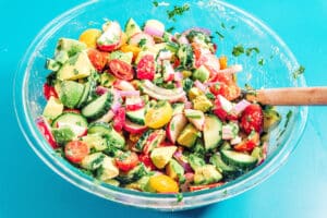 A glass bowl filled with a vibrant avocado salad featuring sliced cucumbers, cherry tomatoes, red onion, and cilantro, with a wooden spoon on a blue background.