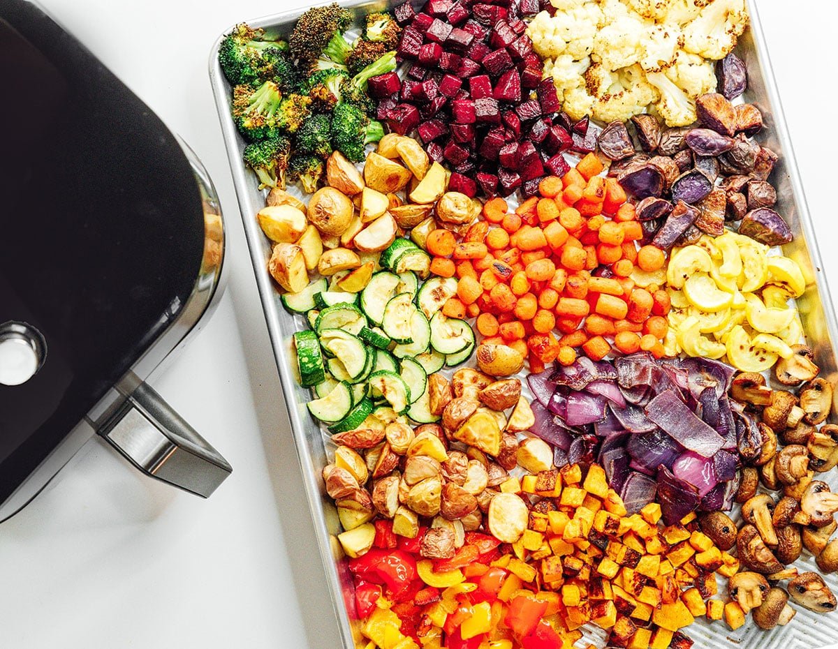 Many colorful veggies on a baking sheet next to an air fryer.