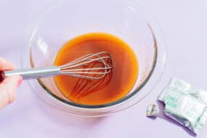 A hand whisking an orange-colored liquid in a glass bowl with ramen noodle salad seasoning packets beside it on a light purple surface.