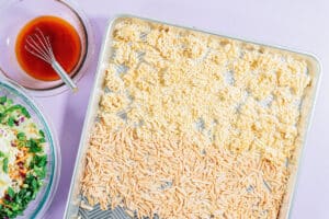 A baking sheet with dry ramen noodles and slivered almonds, set beside a bowl of ramen noodle salad and a bowl of dressing with a small whisk, all arranged on a light purple surface.