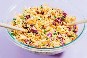 A glass bowl filled with crunchy ramen noodle salad, mixed with shredded cabbage, carrots, green onions, and served with two wooden utensils.