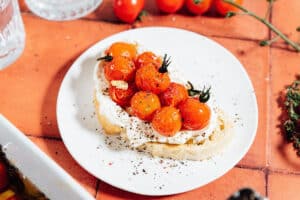A slice of bread topped with whipped cheese, tomato confit, garlic, and cracked black pepper on a white plate.