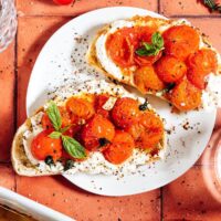 Two slices of toasted bread topped with whipped cheese, roasted cherry tomatoes, fresh basil, black pepper, and a drizzle of infused olive oil on a white plate; cherry tomatoes and herbs nearby on a terracotta surface.
