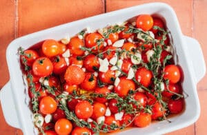 Cherry tomatoes, fresh thyme, chopped garlic, and olive oil in a white baking dish on a terracotta surface—simple ingredients coming together for a flavorful tomato confit, ready to be slow-roasted.