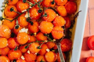 Cherry tomatoes on the vine in a white dish with olive oil, garlic slices, and herbs are transformed into a rich tomato confit, with a spoon scooping up the tender tomatoes.