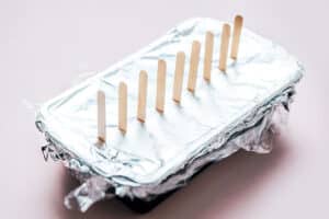 A rectangular loaf pan popsicles container covered with aluminum foil and plastic wrap, with wooden popsicle sticks inserted vertically through the top.