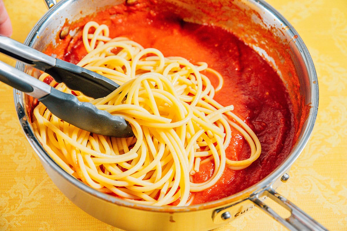 Adding spaghetti noodles to pasta sauce in a pan.