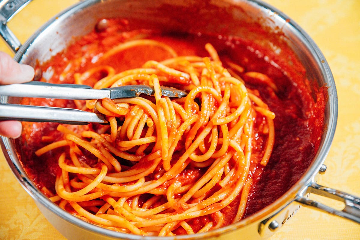 Adding spaghetti noodles to pasta sauce in a pan.