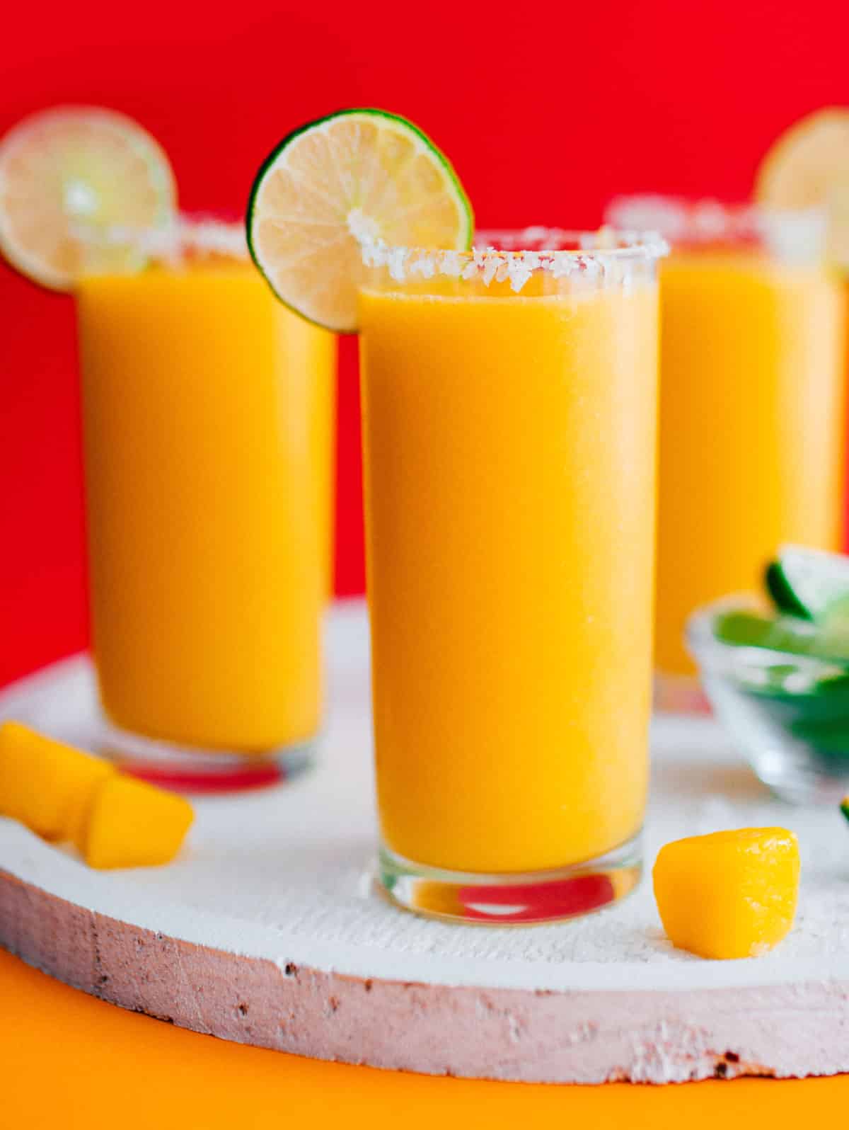 Frozen mango margarita slush in a glass with a slice of lime on a red background.
