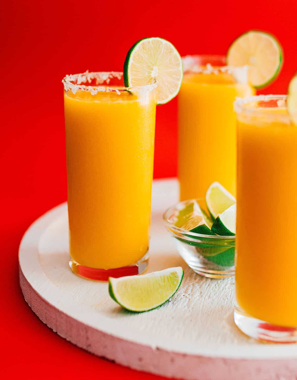 Frozen mango margarita slush in a glass with a slice of lime on a red background.