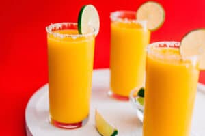 Frozen mango margarita slush in a glass with a slice of lime on a red background.