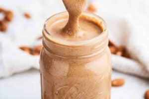 A spoon drizzling creamy almond butter into a glass jar, with whole almonds scattered in the blurred background—perfect for illustrating how to make almond butter at home.