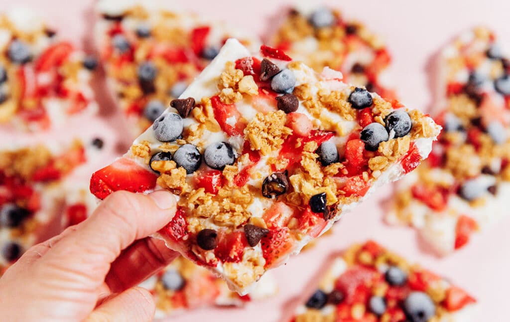 Holding frozen yogurt breakfast bark with berries and granola on a pink background.