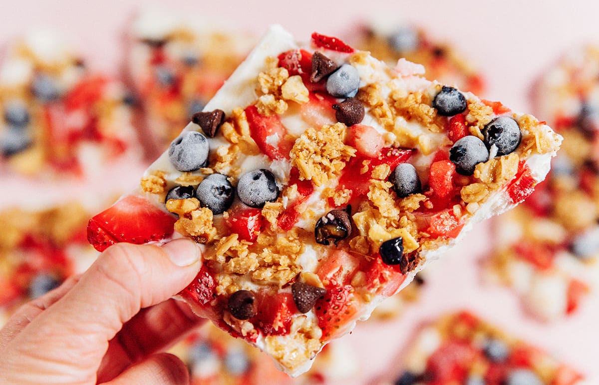 Holding frozen yogurt breakfast bark with berries and granola on a pink background.