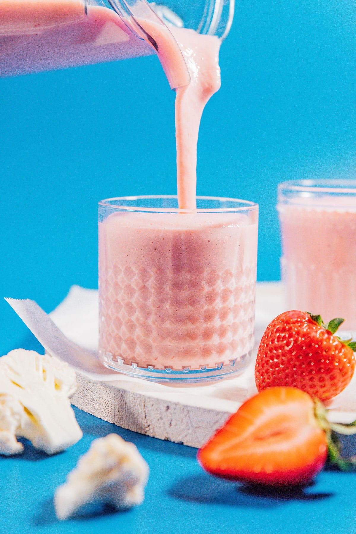 Hidden cauliflower smoothie with strawberry and paper straws in a glass on a blue background.