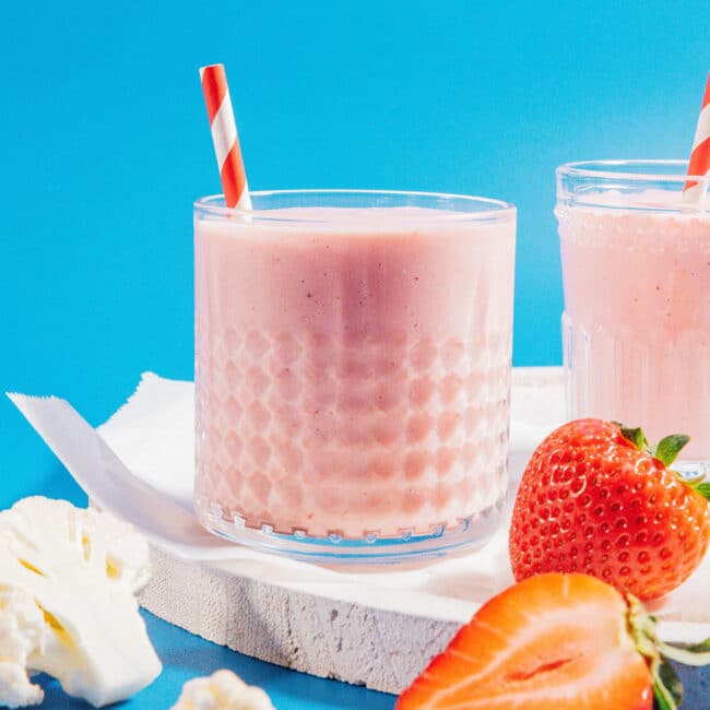 Hidden cauliflower smoothie with strawberry and paper straws in a glass on a blue background.