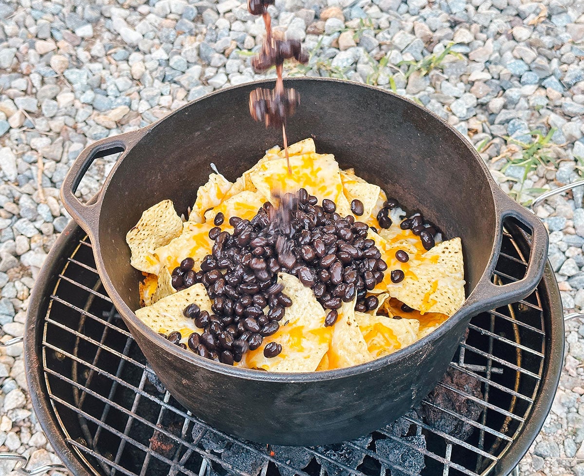 Adding beans to nachos in a Dutch oven over a coal stove.