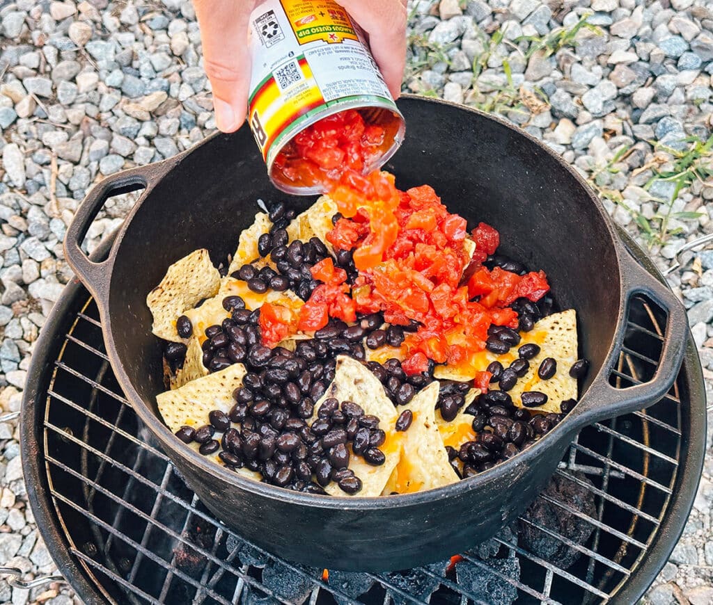 Adding tomatoes to nachos in a Dutch oven over a coal stove.