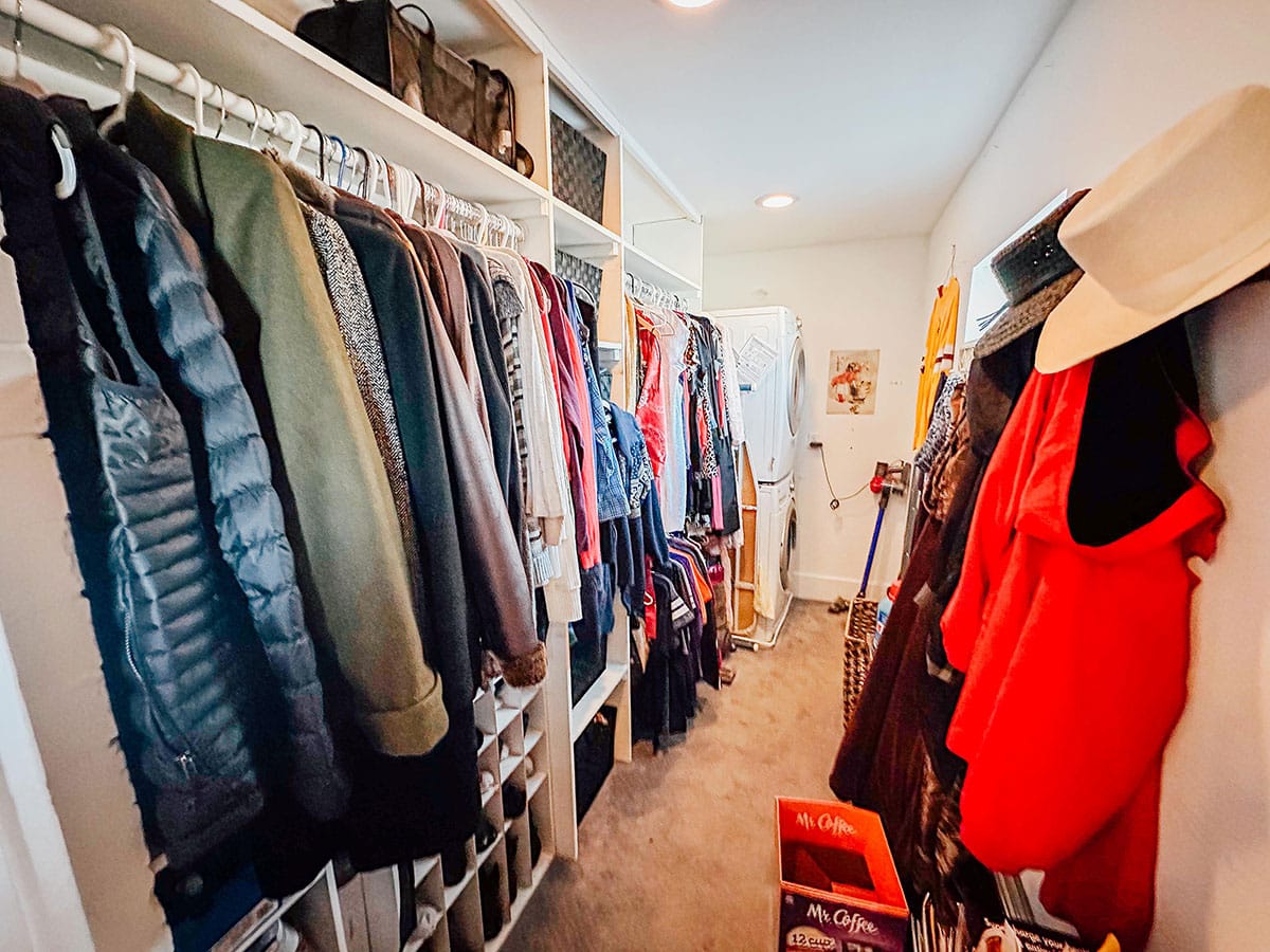 A walk-in closet with clothes hanging on both sides, hats on hooks, bags on shelves, and boxes labeled Mr. Coffee on the carpeted floor.