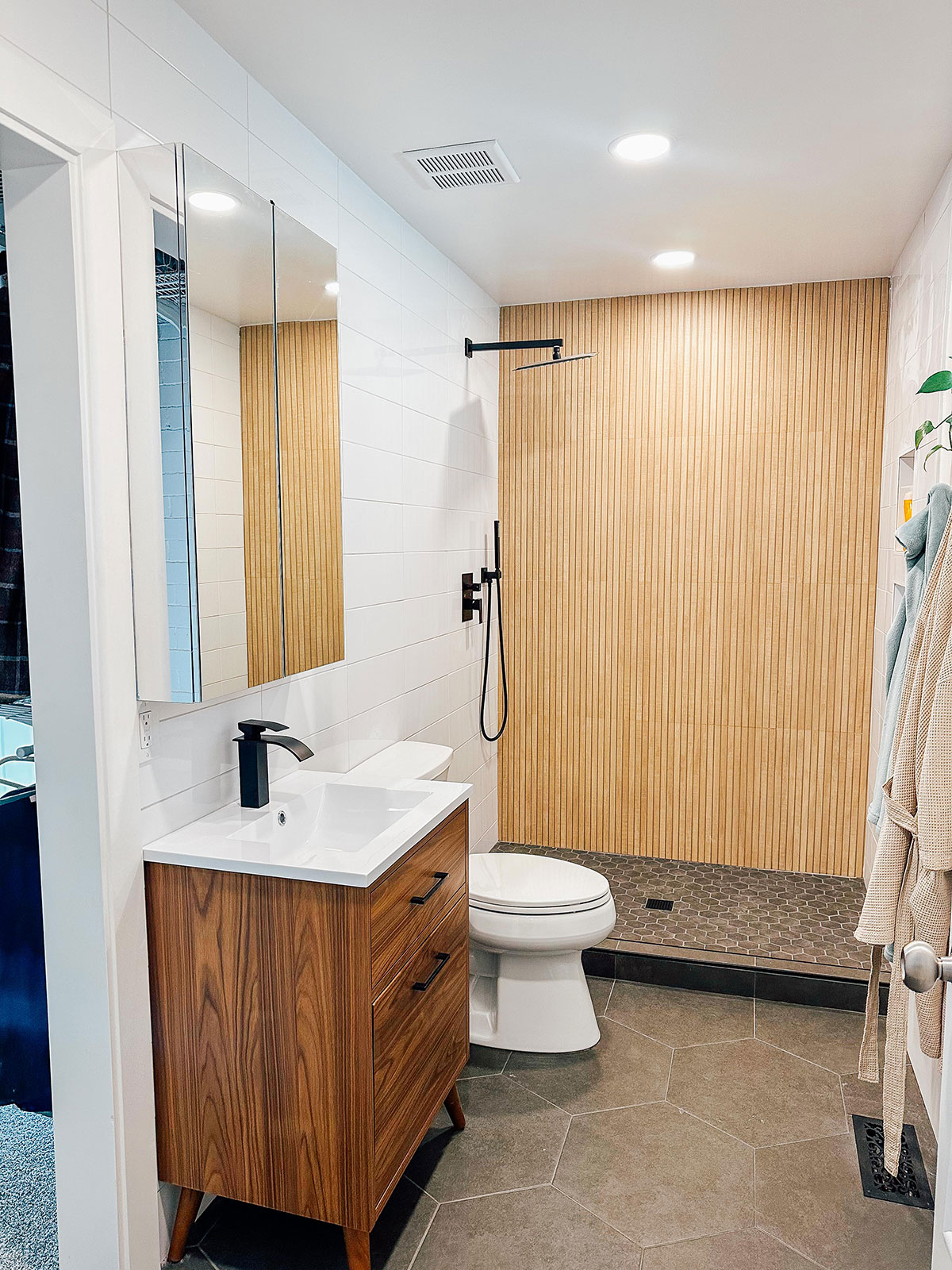 Modern bathroom with wood vanity, white sink, wall-mounted mirror, toilet, walk-in shower with wood accent wall, and hexagonal floor tiles.
