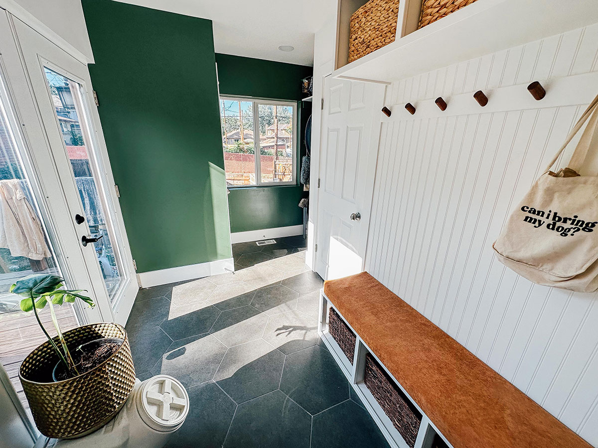 A bright mudroom with green and white walls, a bench with storage baskets, coat hooks, a tote bag, a potted plant, and a door leading outside.
