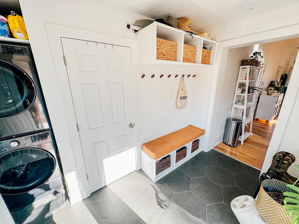 A laundry room with a stacked washer and dryer, a white bench with cubbies and baskets, coat hooks, overhead storage, and a door leading to another room with wood flooring.