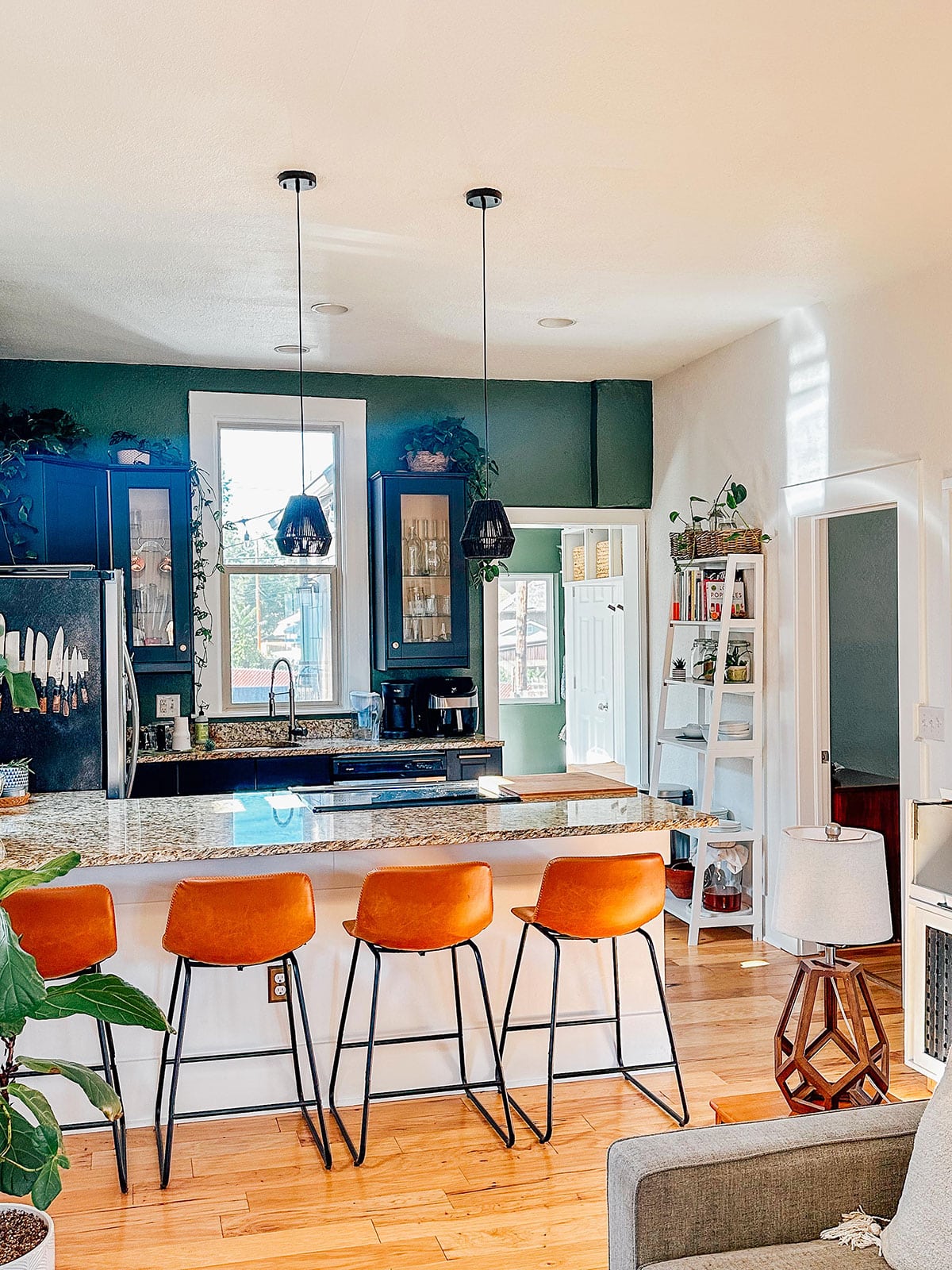 Modern kitchen with green accent wall, black cabinets, granite island, and four orange bar stools; pendant lights hang over the island.