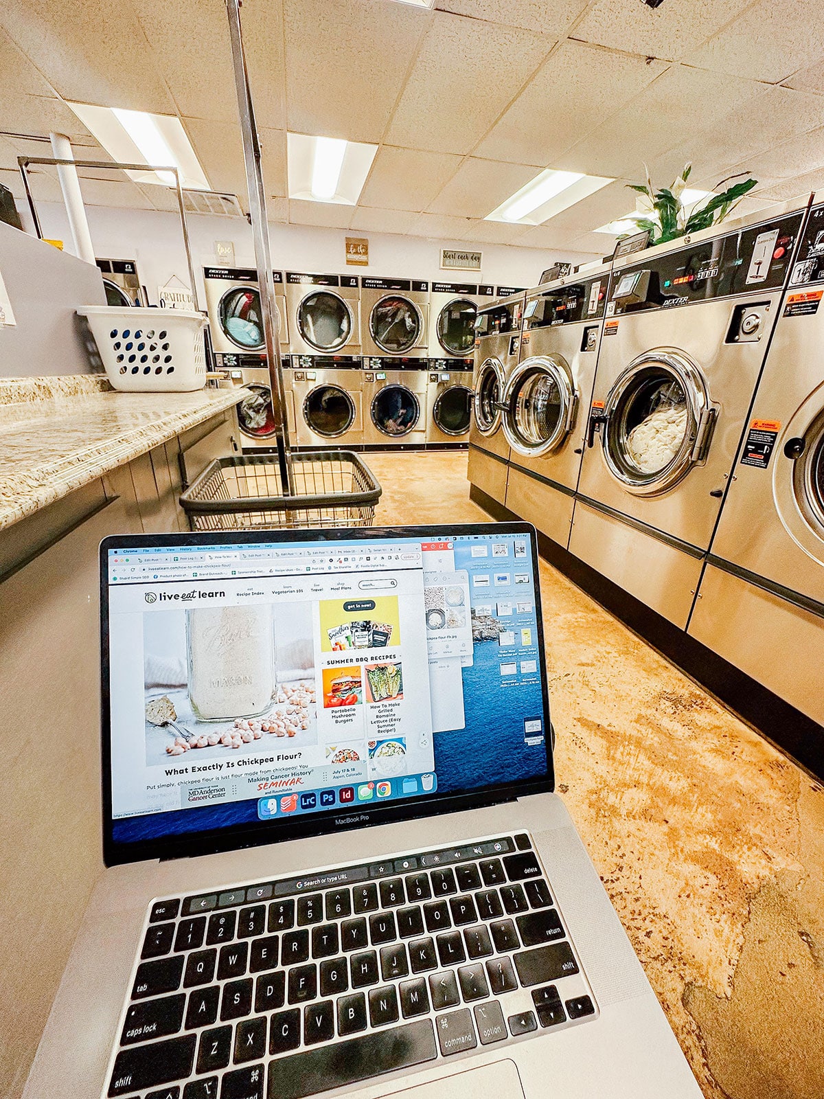 A laptop displaying a website is open on a counter in a laundromat with multiple front-loading washing machines in the background.