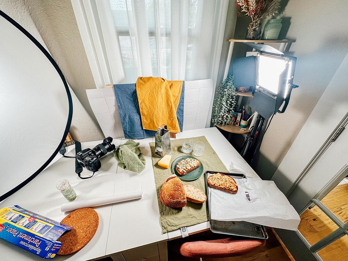 A tabletop setup with bread, cheese, and kitchen items arranged for food photography, surrounded by camera equipment, lighting, and props near a window.