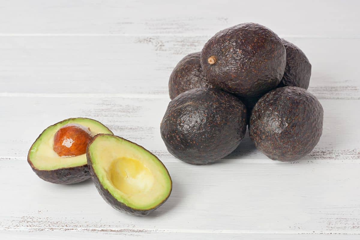 a stack of stewart avocados next to one cut open on a white wood table.