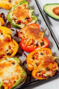 Colorful bell pepper halves—green, red, and yellow—are filled with a savory quinoa stuffed peppers blend of rice, beans, and cheese, then baked to perfection. Served on a tray alongside a fresh halved avocado.