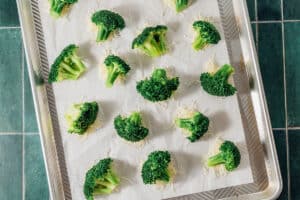Smashed broccoli florets placed on parchment paper atop a metal baking sheet, ready for roasting, on a green tiled surface.