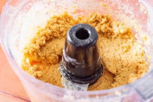 A food processor bowl containing thick, mixed chickpea pizza crust dough with a central black blade attachment.