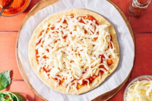 Unbaked chickpea pizza crust on parchment paper with tomato sauce and shredded cheese, placed on a round baking tray with ingredients visible around it.