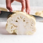 A hand demonstrates how to cut cauliflower, skillfully slicing through a head of cauliflower on a white surface.
