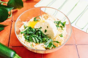 A clear glass bowl contains shredded cheese, a cracked egg, ricotta cheese, and chopped fresh herbs on a tiled countertop with sliced zucchini nearby.