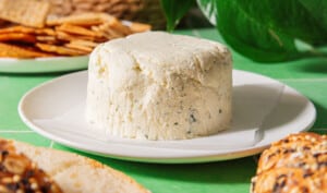 A round block of herbed vegan cheese sits on a white plate, surrounded by slices of bread and crackers on a green surface.