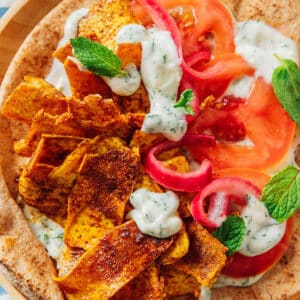 A pita topped with seasoned, roasted slices, fresh tomato, red onion, dollops of tzatziki, and mint leaves, served on a wooden plate.