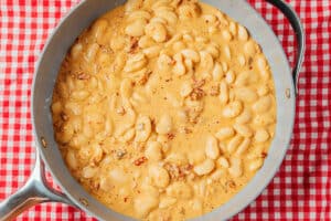 A skillet filled with creamy cooked lima beans and pieces of bacon sits on a red and white checkered tablecloth.