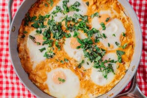 A skillet with eggs cooked in a tomato-based sauce, topped with chopped fresh herbs, placed on a red and white checkered cloth.