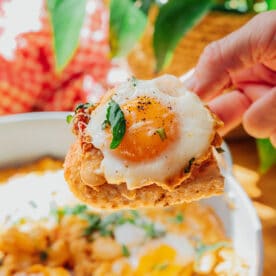 A hand holds up a piece of toast topped with a fried egg, fresh basil, and black pepper over a plate of similar toasts, with a green plant in the background.