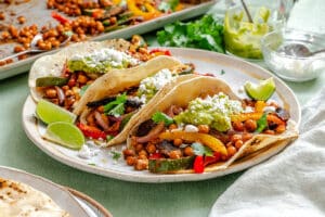 Three soft tacos filled with roasted chickpeas, bell peppers, zucchini, guacamole, cilantro, and crumbled cheese on a plate with lime wedges.