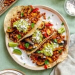 Three soft tacos filled with roasted chickpeas, sautéed vegetables, guacamole, fresh cilantro, and crumbled cheese, served with lime wedges on a white plate.