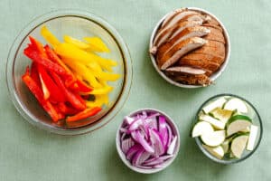 Four bowls containing sliced red and yellow bell peppers, sliced mushrooms, sliced red onions, and chopped zucchini on a green surface.