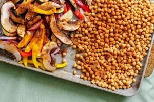 A sheet pan with sliced mushrooms, bell peppers, red onions, and seasoned chickpeas, ready to be roasted.